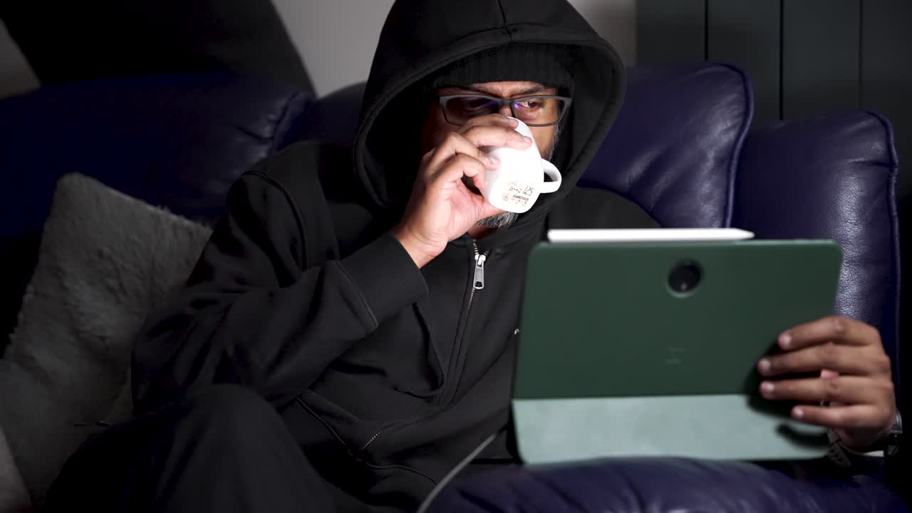 South Asian Man In Hoodie Using Tablet With Stylus Pen On Sofa At Home Relaxing And Browsing Online Watching Videos Or Video Conferencing