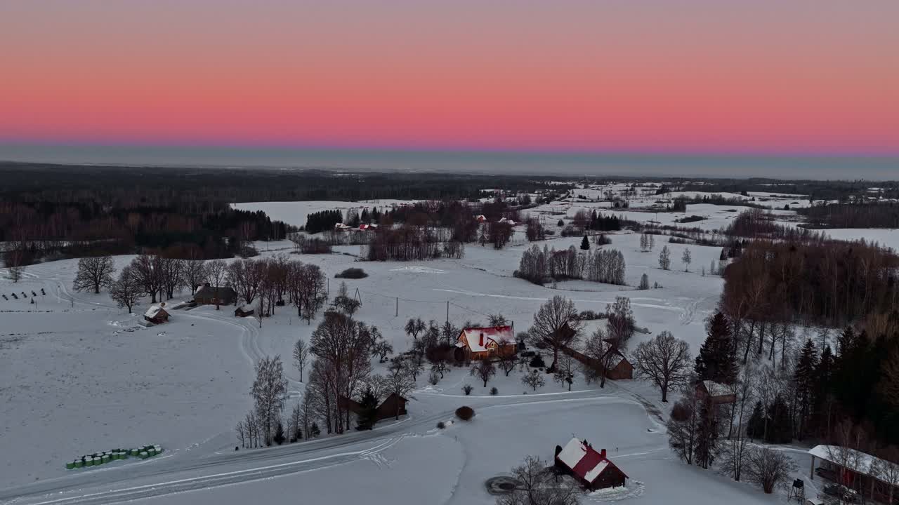 Dronen ilmakuva lumipeitteisen viljelysmaan ja pienen kylän yli vaaleanpunaisen auringonlaskun taivaan alla, rauhallista talvista maaseutua tyhjää tilaa tekstille.