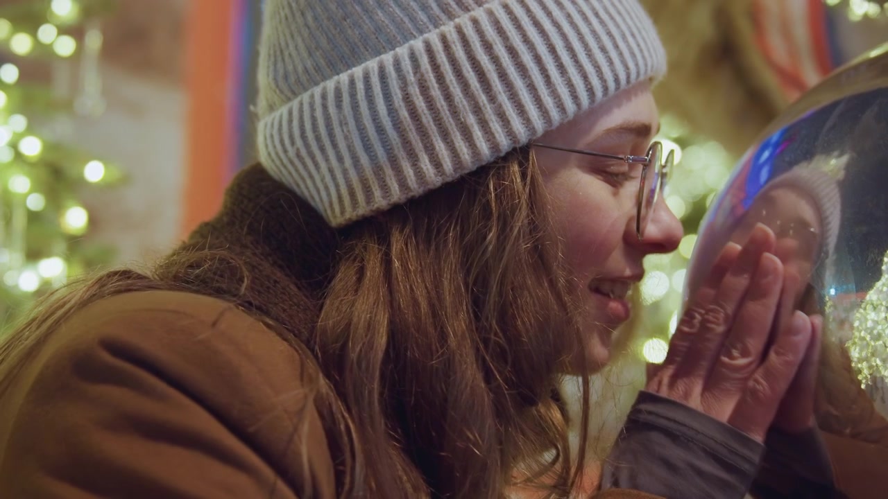 Donna caucasica che si riflette in una sfera lucida, bagliore accogliente del mercato con luci bokeh calde, indossa un berretto grigio e occhiali, cappotto marrone, sorriso delicato e sguardo contemplativo, mani vicino alla sfera, atmosfera notturna intima.