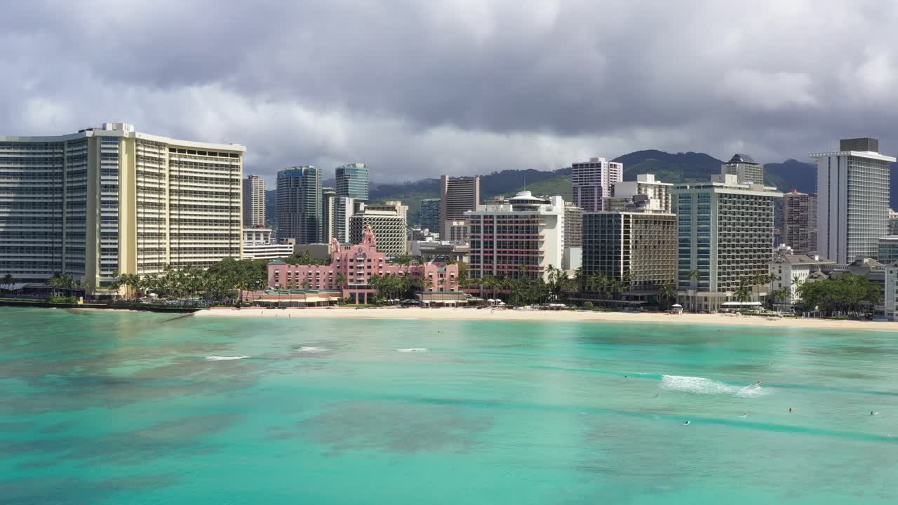 Drone video capturing the vibrant turquoise waters of Waikiki Beach against the stunning Honolulu skyline. Dramatic clouds and diverse architecture create an enchanting, picturesque scene