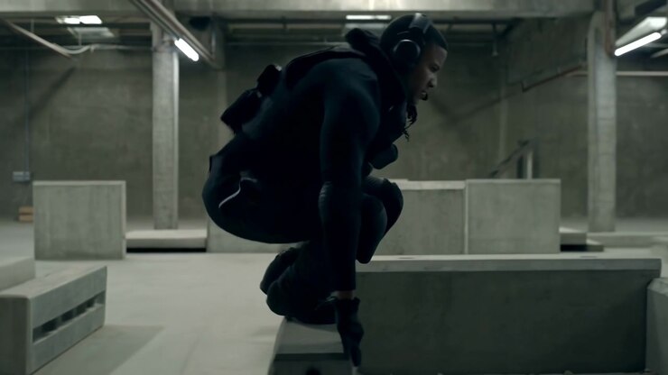 A man in dark tactical clothing stands in profile inside a large industrial training hall with concrete walls, overhead pipes, and block barriers. The muted