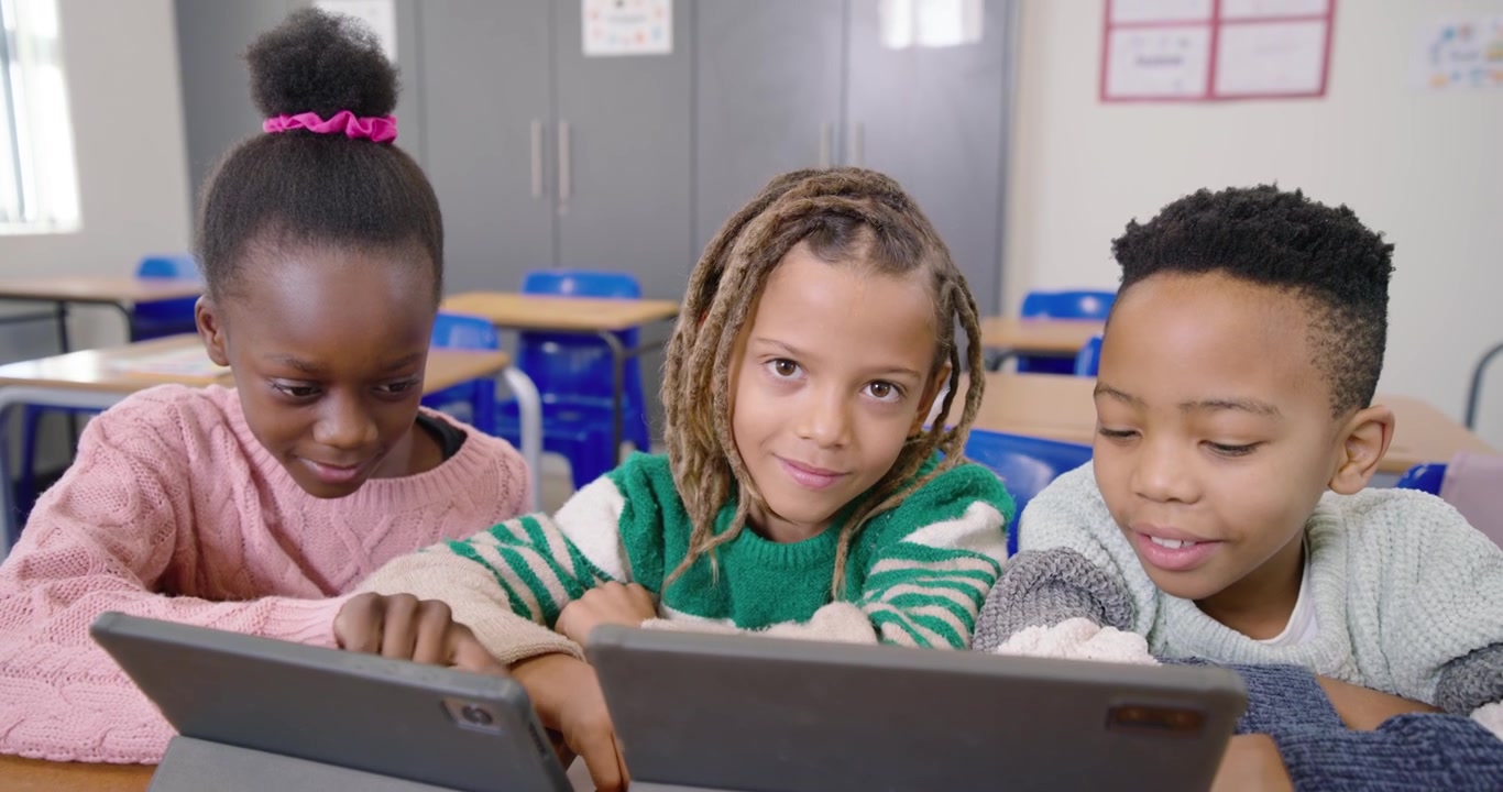 Studenti delle scuole elementari che osservano due tablet, toccandoli e appoggiandosi alla scrivania, mentre collaborano al lavoro scolastico