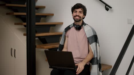 Hombre-Barbudo-Sonriente-Con-Computadora-Portátil,-Sentado-En-Escaleras-De-Madera-En-Casa-En-Cuadros