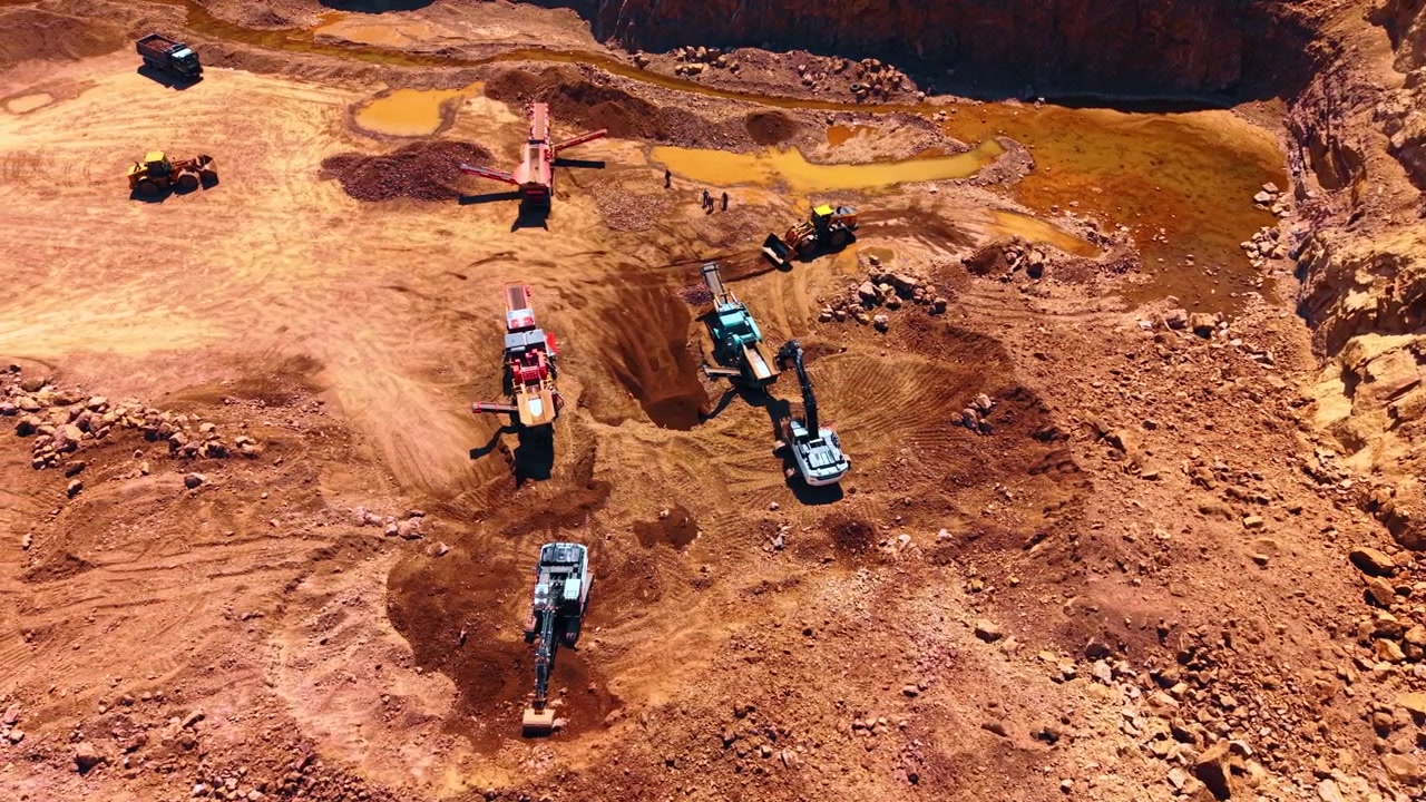 Aerial view of heavy machinery operating in open pit rock quarry. Excavators and crushing machines process extracted stones on reddish sandy soil under bright daylight.