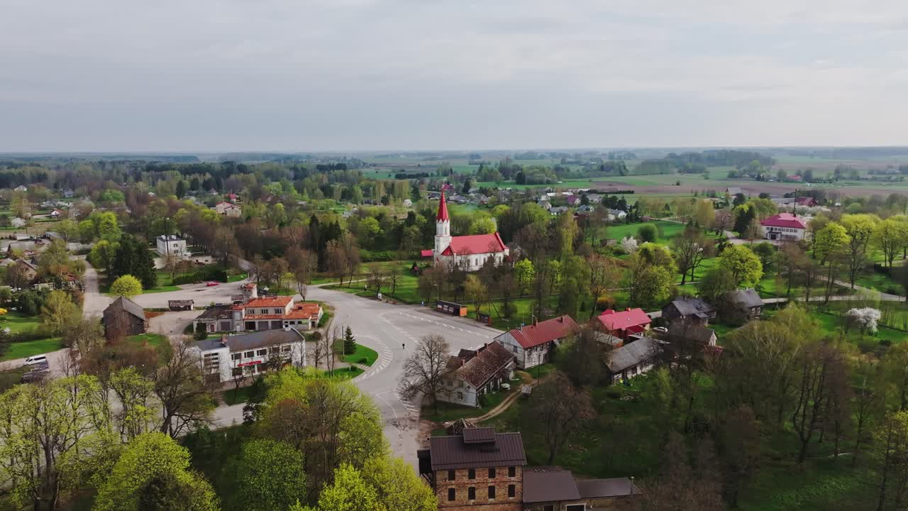 Drone pulls back from Rucava Lutheran Church surrounded by green trees, Latvia