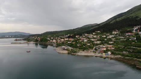 drone view in albania flying over shkodër lake in pogradec on cloudy day circling a road with houses looking at the lake and mountain behind