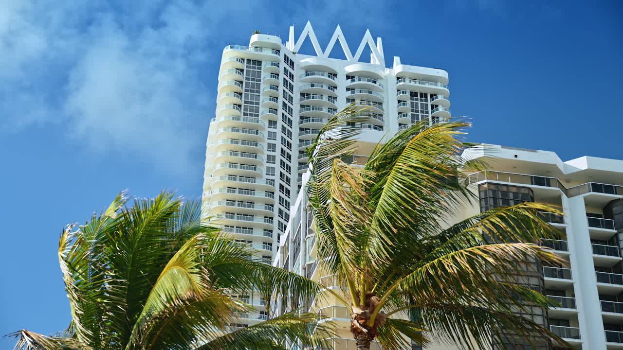 Grattacielo moderno con architettura a corona bianca a Miami. Alto edificio residenziale con balconi curvi e corona decorativa bianca contro il cielo blu e foglie di palma.