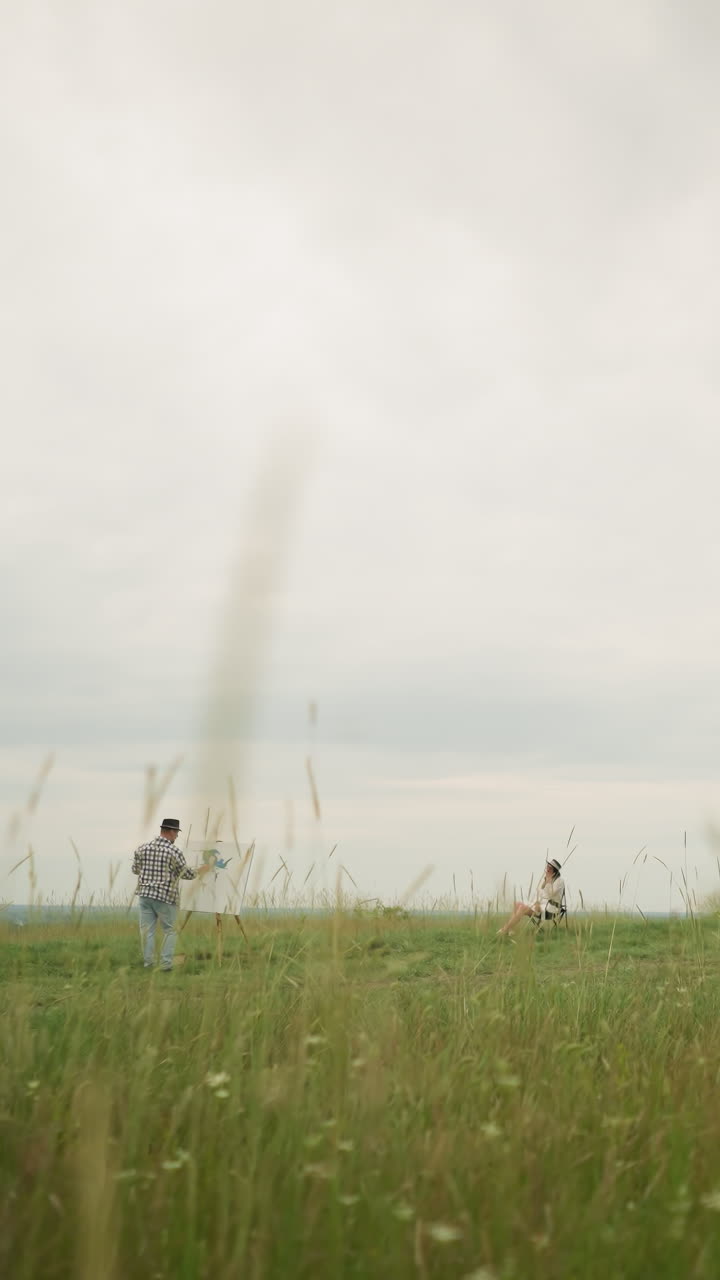 한 화가, 모자와 체크 셔츠를 입고, 높은 잔디로 가득 찬 초원에서, 구름이 가득 찬 하늘 아래에 easel에 그림을 그리고 있습니다. 모자와  옷을 입은 여자가 의자에 평화롭게 앉아 있습니다.