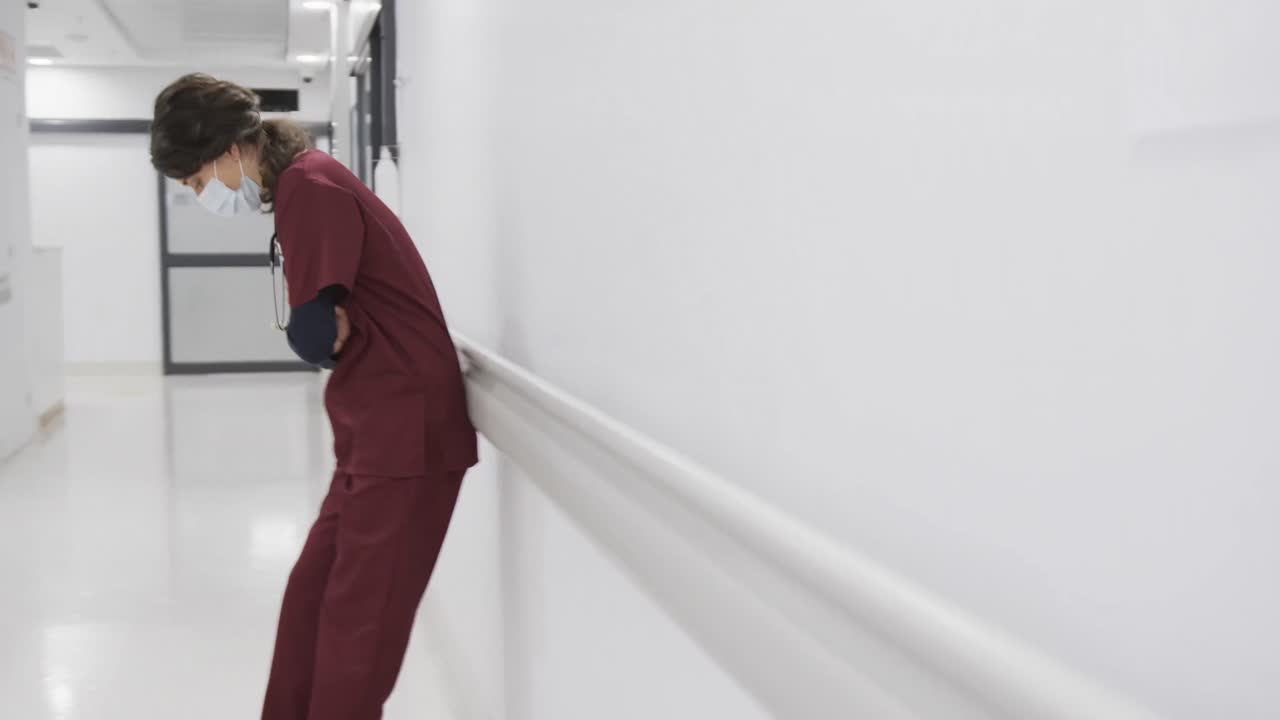 doctora caucásica cansada con máscara facial sentada en el pasillo del hospital, cámara lenta