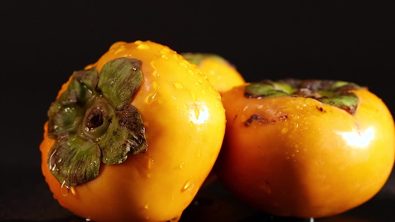 Detailed view of water droplets on vibrant persimmons with green calyx, set against a dark backdrop.