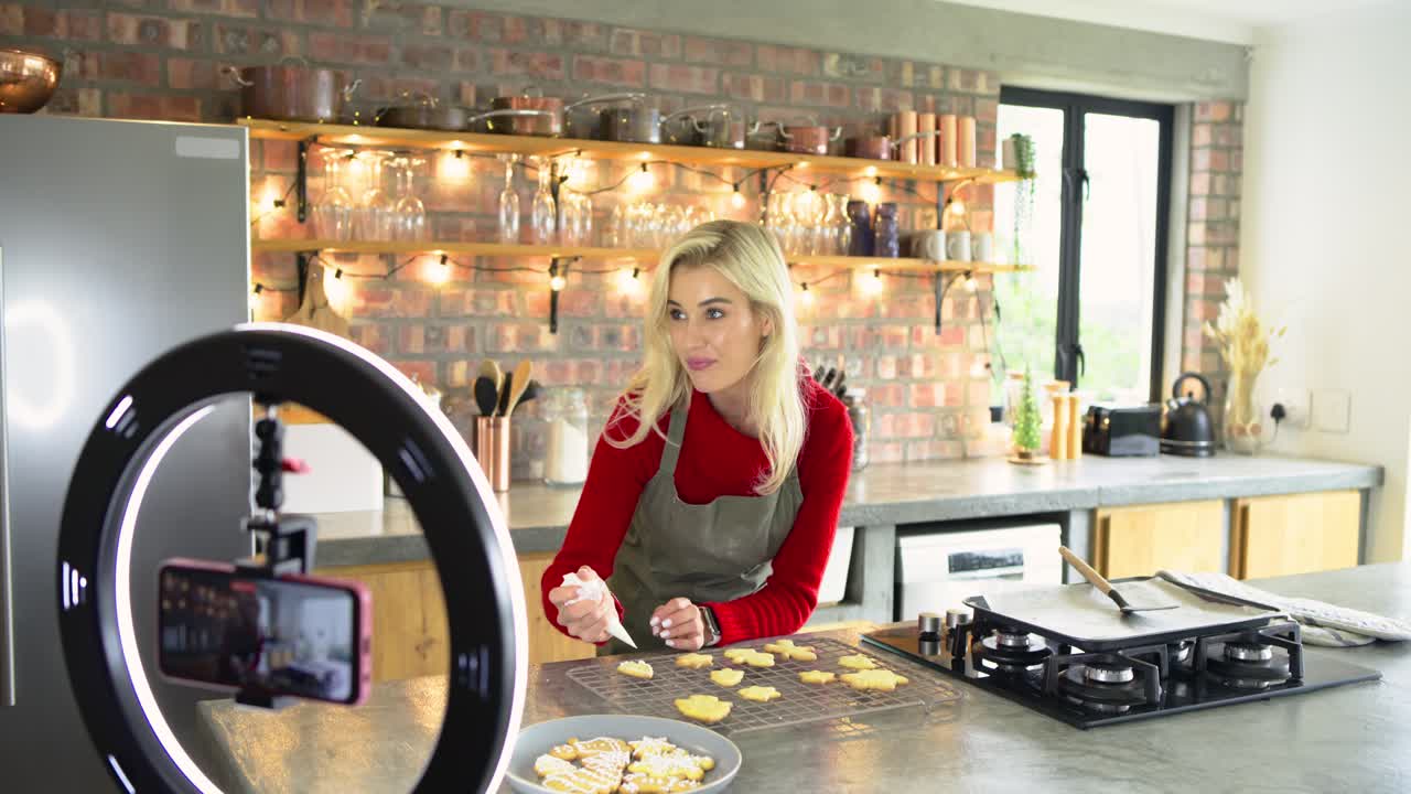 Female baker squeezing icing onto shaped cookies at island with ring light, smartphone filming