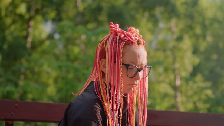 Persona con capelli colorati assorta nella lettura, persona con trecce vivaci che studia in silenzio immersa nella natura, studente con capelli rosa acceso seduto pensieroso su una panchina tra le foglie al tramonto