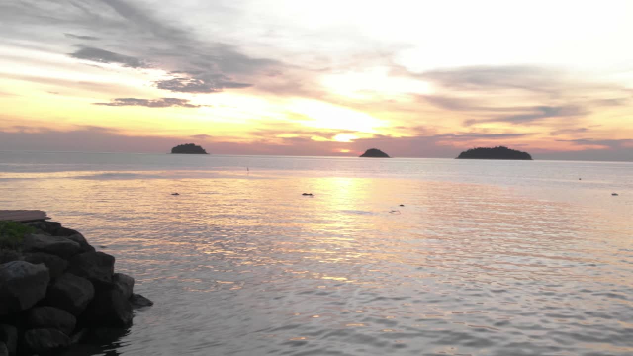 Aerial drone shot moving from a narrow canal into the open ocean with the sun setting on the horizon, capturing warm golden light and calm waters