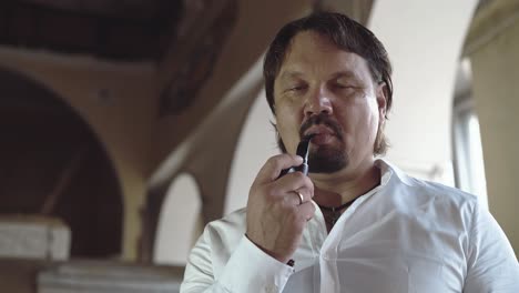 Portrait-of-a-brutal-young-man-with-a-beard-that-smokes-a-pipe-then-offers-it-to-the-viewer-smiling-a-close-up