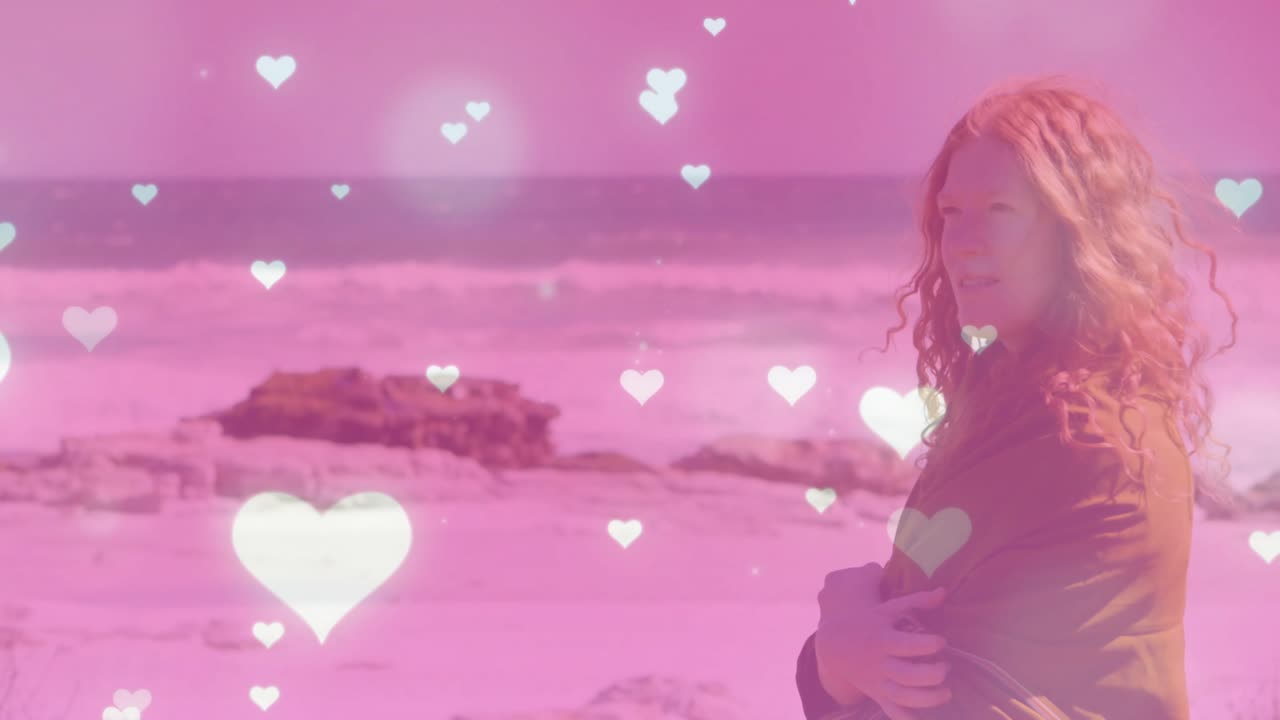 Donna che si avvolge nel cappotto sulla riva, mentre la brezza le solleva i capelli e cuori rosa si moltiplicano per marketing.
