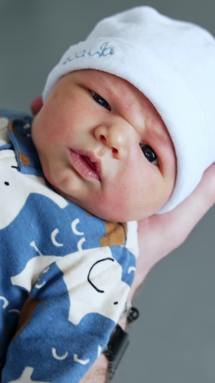 Cute dark-eyed newborn boy in blue romper and white cap. Few hours old child in dad's hands. Vertical video
