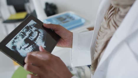 Biracial-female-dentist-examining-teeth-on-tablet-at-modern-dental-clinic