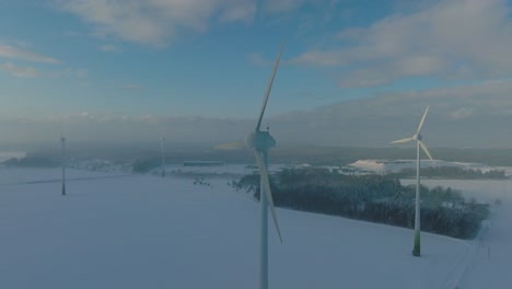 aerial view of wind turbines generating renewable energy in the wind farm, snow filled countryside landscape with fog, sunny winter day with some clouds, wide orbiting drone shot