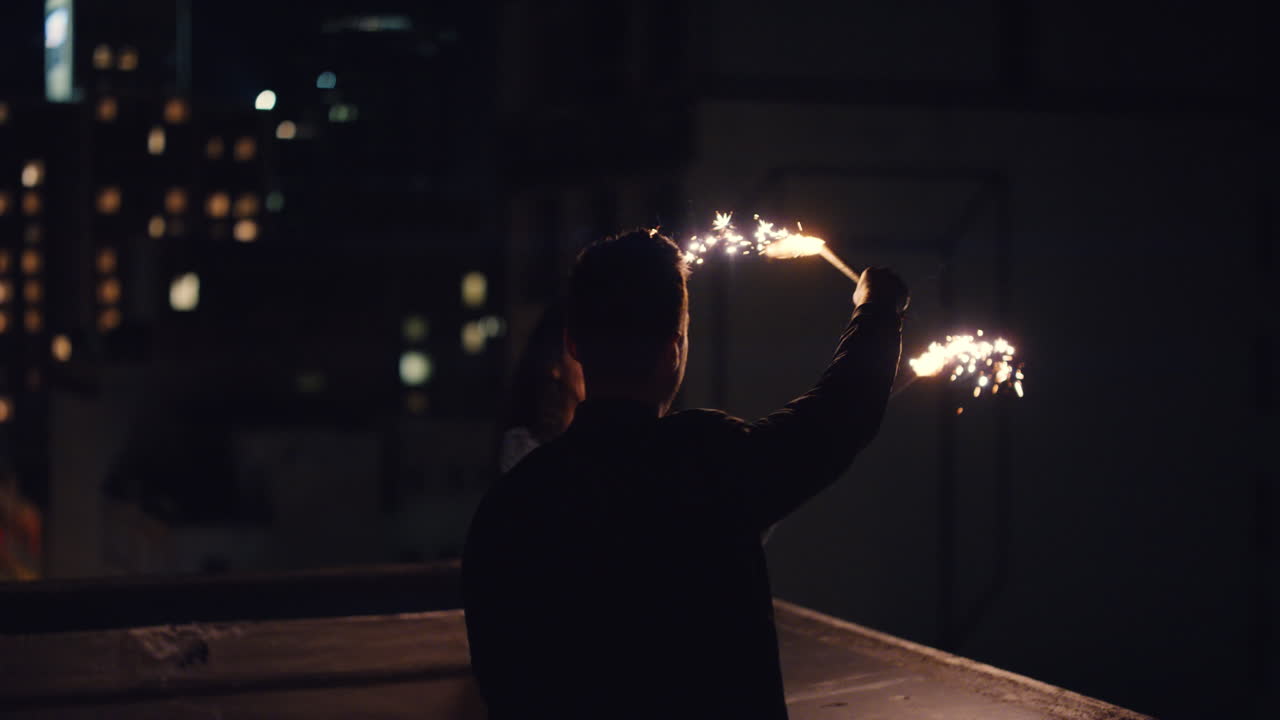 feliz pareja caucásica besándose en la azotea por la noche sosteniendo bengalas celebrando el aniversario disfrutando de la romántica noche urbana de nochevieja