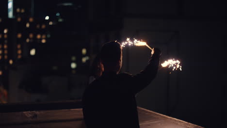 happy-caucasian-couple-kissing-on-rooftop-at-night-holding-sparklers-celebrating-anniversary-enjoying-new-years-eve-romantic-urban-evening