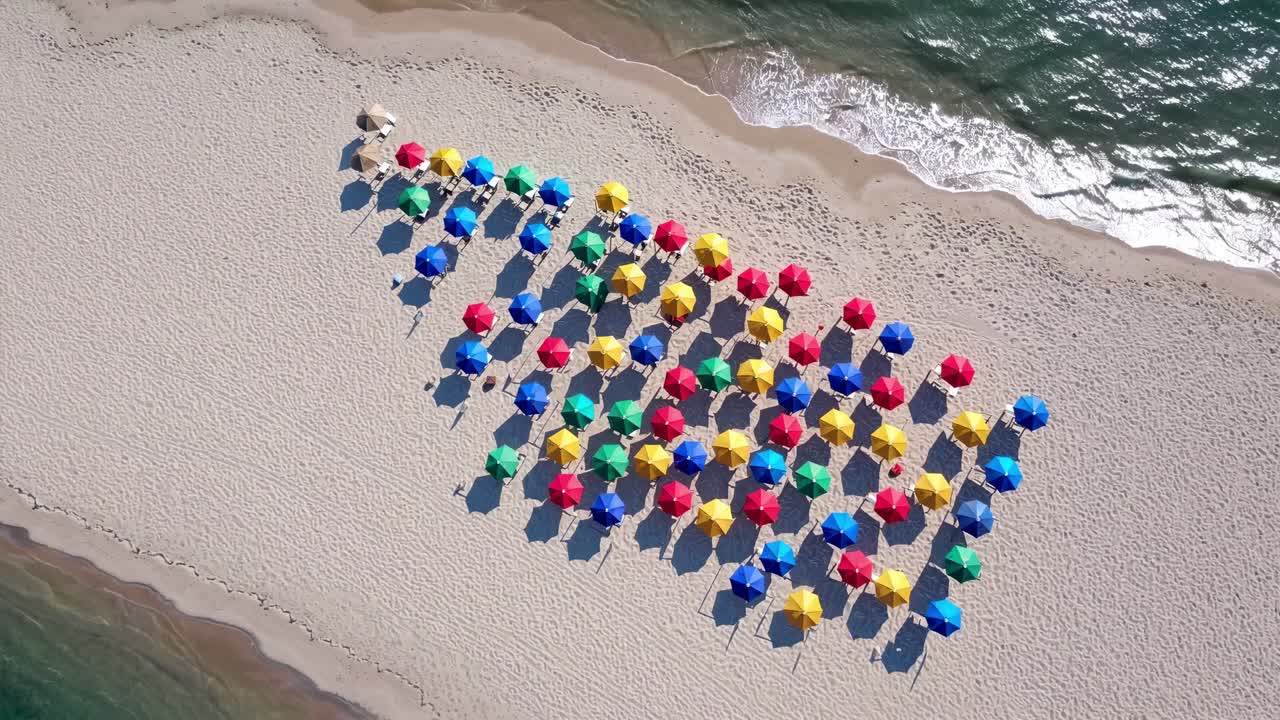Aerial view revealing a vibrant beach scene filled with colorful umbrellas in a striking pattern, contrasting against white sand and gently lapping blue ocean waves