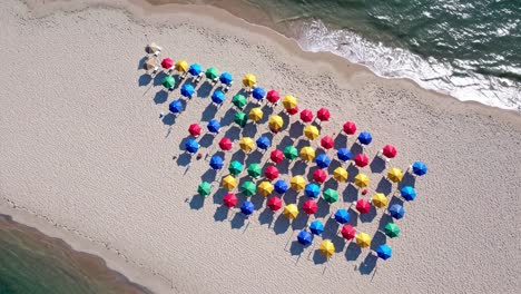 aerial view revealing a vibrant beach scene filled with colorful umbrellas in a striking pattern, contrasting against white sand and gently lapping blue ocean waves