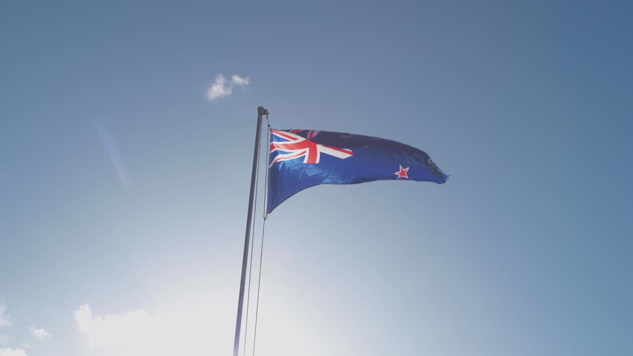 una bandera de nueva zelanda ondea en un poste con cielo azul y bengalas solares