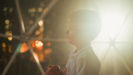 sleepy child stands against lighting near transparent tent wall. dreamy blond kid twirls hands thoughtfully in evening closeup on blurred background slow motion