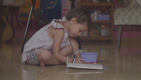 una linda niña sentada en el piso de una sala de estar dibujando y coloreando imágenes en su libro de colorear en casa adentro