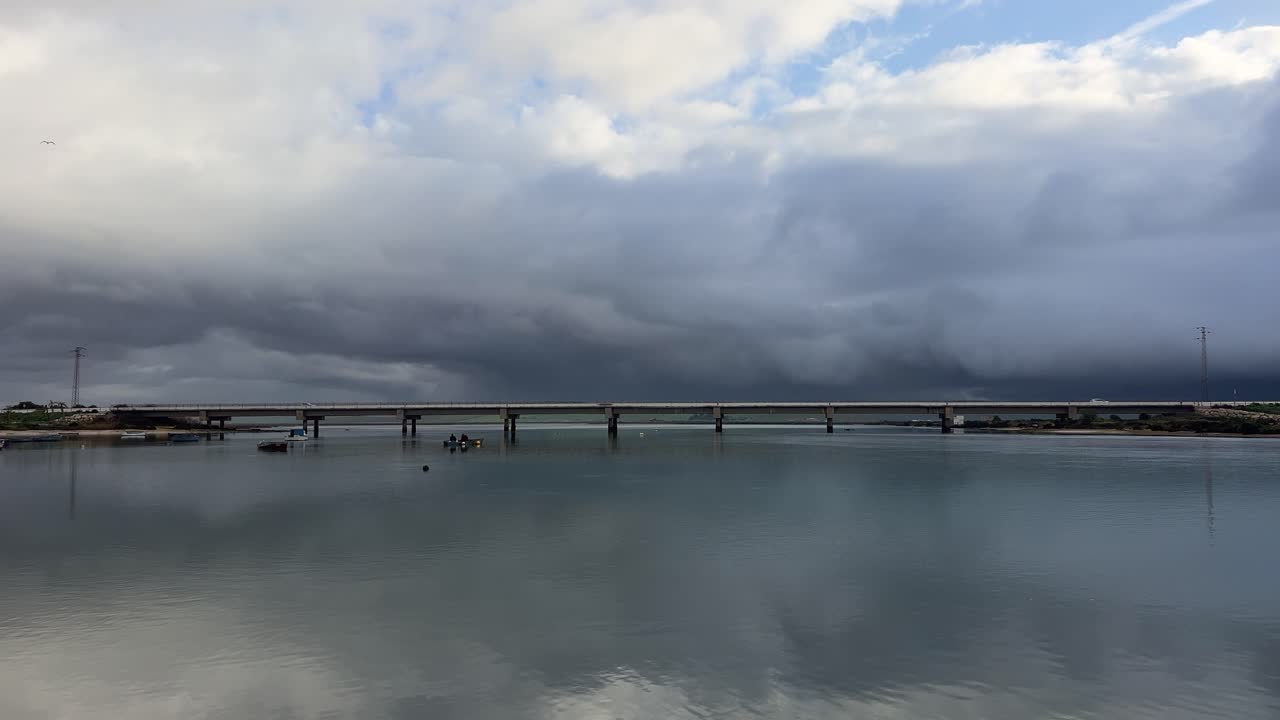 Stormy skies loom over a calm river in Barbate, Andalusia, with a bridge stretching across the water. The scene captures the contrast between the dark clouds and the serene reflection