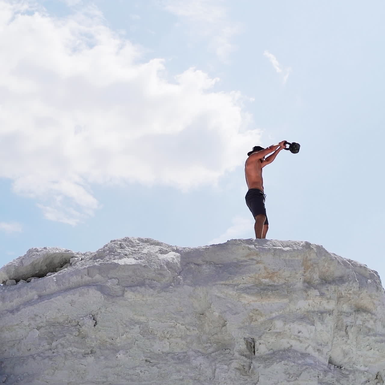Side view of a strong man lifting kettlebell on white hill. Male athlete swinging kettlebell with two hands on the beautiful natural background outdoors.