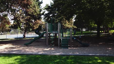 Leerer-Spielplatz-Mit-Beschilderung,-Die-Kinder-Verbietet,-Und-Lockdown-Covid-19-Beschränkungen-Der-Sozialen-Distanzierungspandemie-In-Einem-üppig-Grünen-Sommerpark-Mit-Aktiven-Menschen,-Die-Im-Hintergrund-Tennis-Spielen-1-2