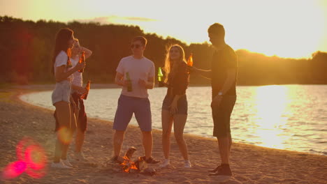 the five young people spend time at sunset on the beach in shorts and t-shirts around bonfire with beer. they are dancing and enjoying the warm summer evening near the river.