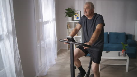 athletic middle-aged man is training on exercycle in living room healthy lifestyle at isolation period during pandemic keeping good physical condition at middle age