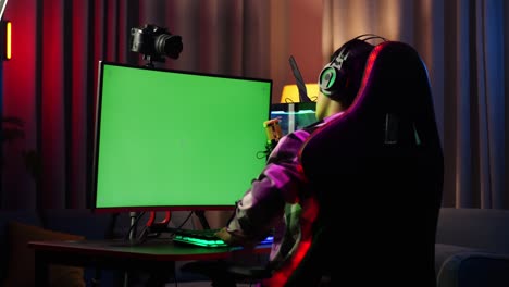 back view of asian boy streamer having a backache while using green screen personal computer. live stream on desk illuminated by rgb led strip light