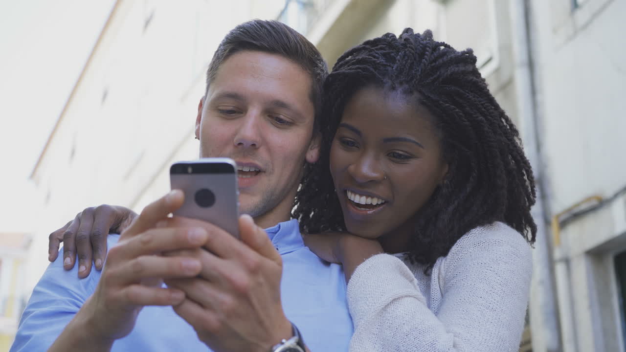 una pareja multirracial sonriente con teléfono inteligente al aire libre.