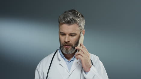 male physician speaking on the mobile phone and doing consultation by connection on the gray wall background