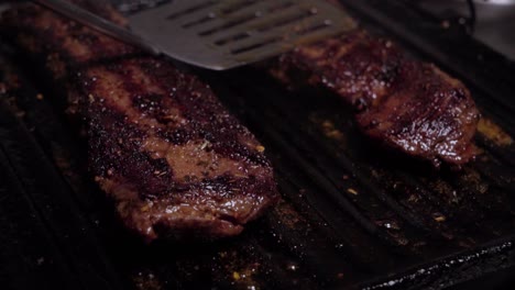 spatula pressing down on two beef steaks grilling on cast iron griddle, close up