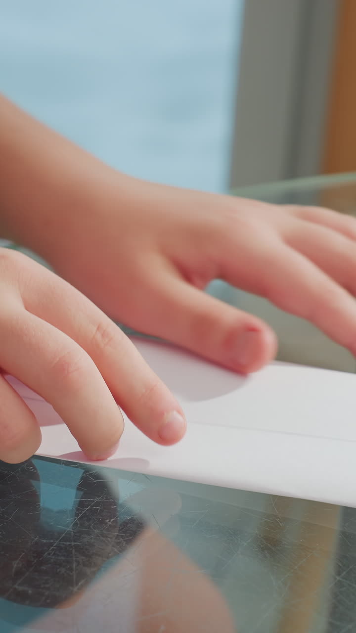 Kids craft airplane, Child shaping paper cone, Child carefully folds paper on glass table, Young child gently folds paper cone on glass surface with sunlight filtering through window