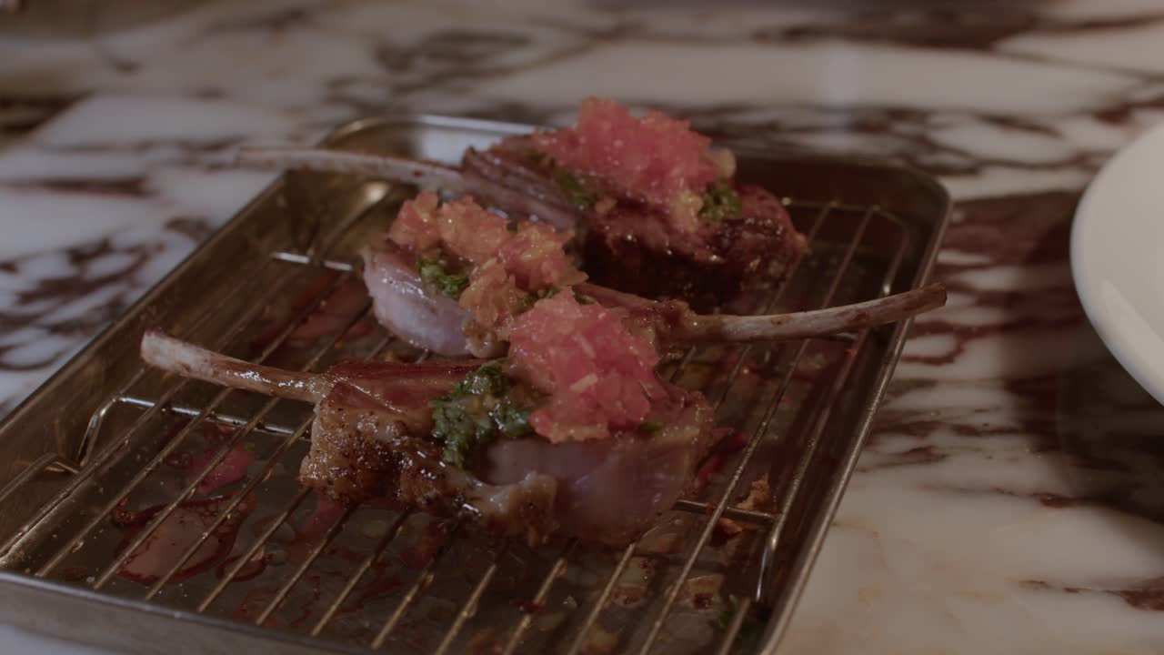 A detailed close-up shot of three perfectly grilled lamb chops resting on a metal cooling rack, topped with a vibrant green herb sauce and fresh tomato salsa