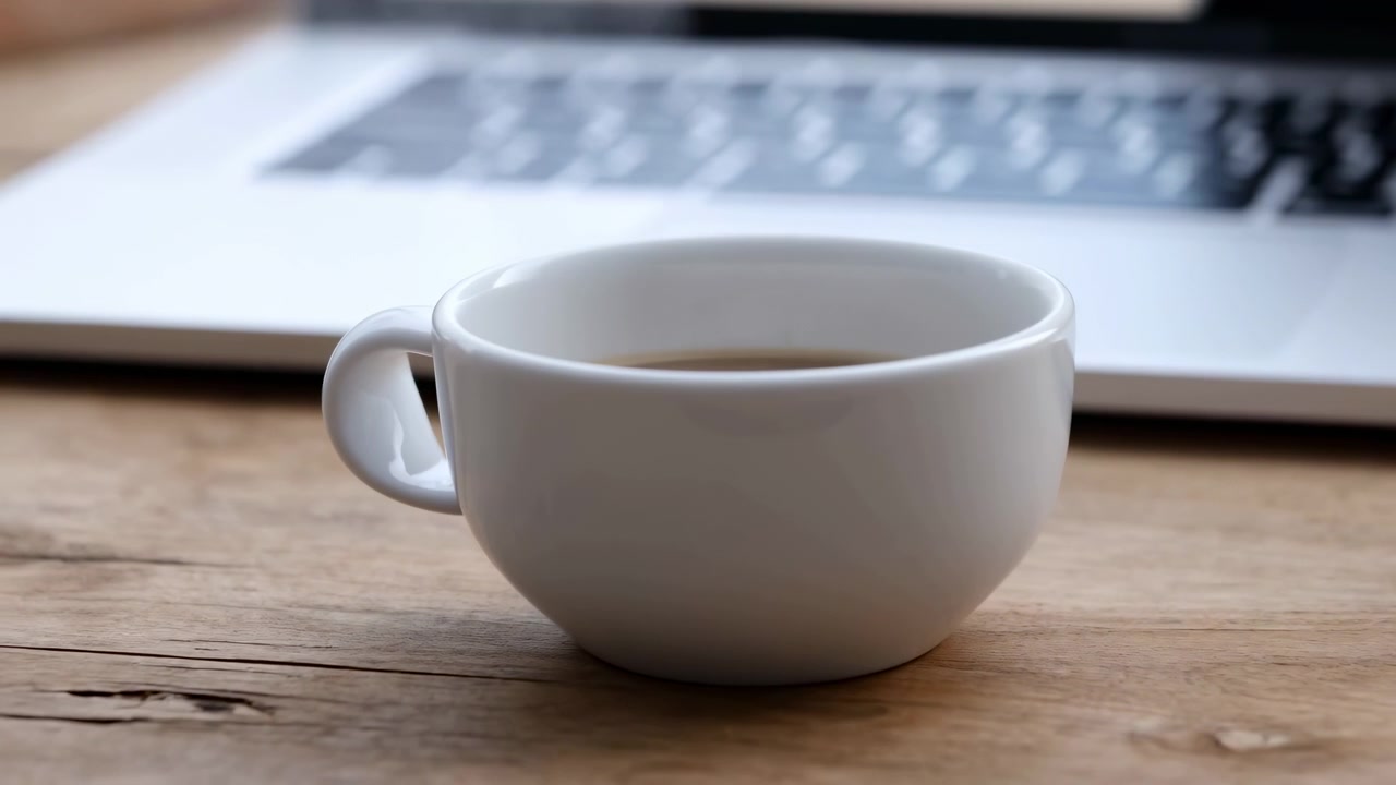 Primo piano a metà sequenza di una tazza di caffè in ceramica bianca su una scrivania di legno durante una pausa di lavoro con un laptop.