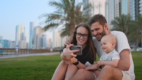 familia feliz con dos hijos sentados juntos en la hierba en el parque y tomando una selfie. con teléfono inteligente.