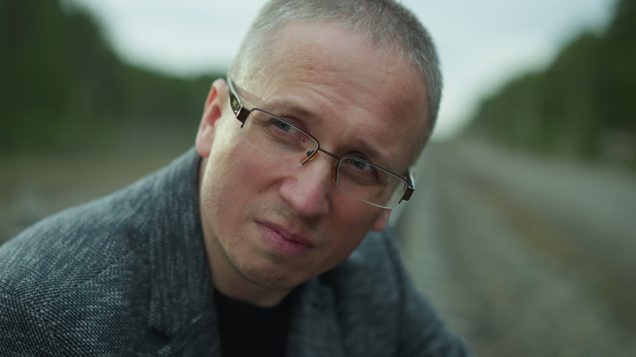 심각한 표정으로 안경을 입은 중년 남자의 근접한 모습, 그는 회색 재을 입고 나무의 흐릿한 배경으로 야외에 있습니다.