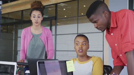 Three-happy-diverse-creative-male-and-female-colleagues-using-laptops-talking-in-modern-interior