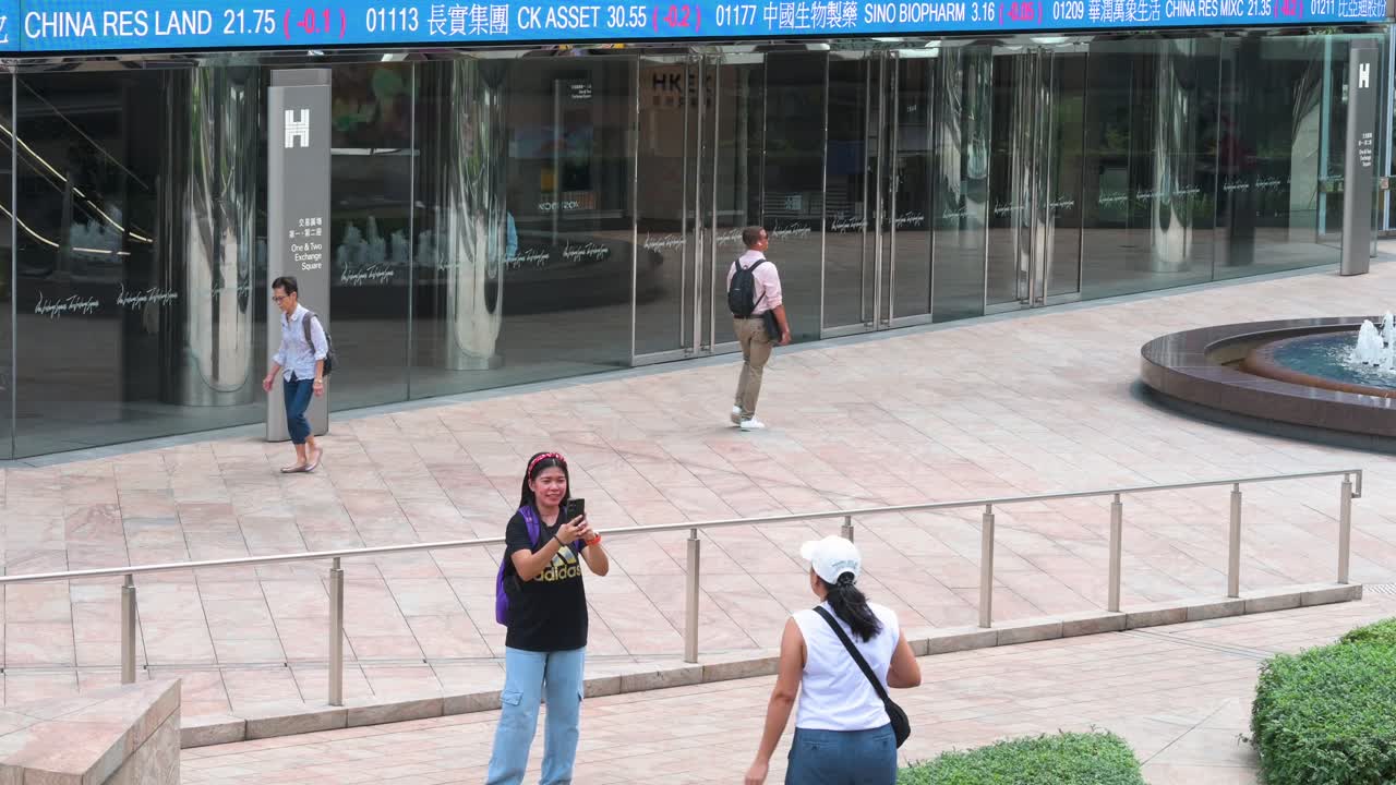 As people take photos, a moving screen in the background displays negative stock ticker symbols at Exchange Square, home of the Hong Kong Stock Exchange, in Hong Kong's financial district, China.