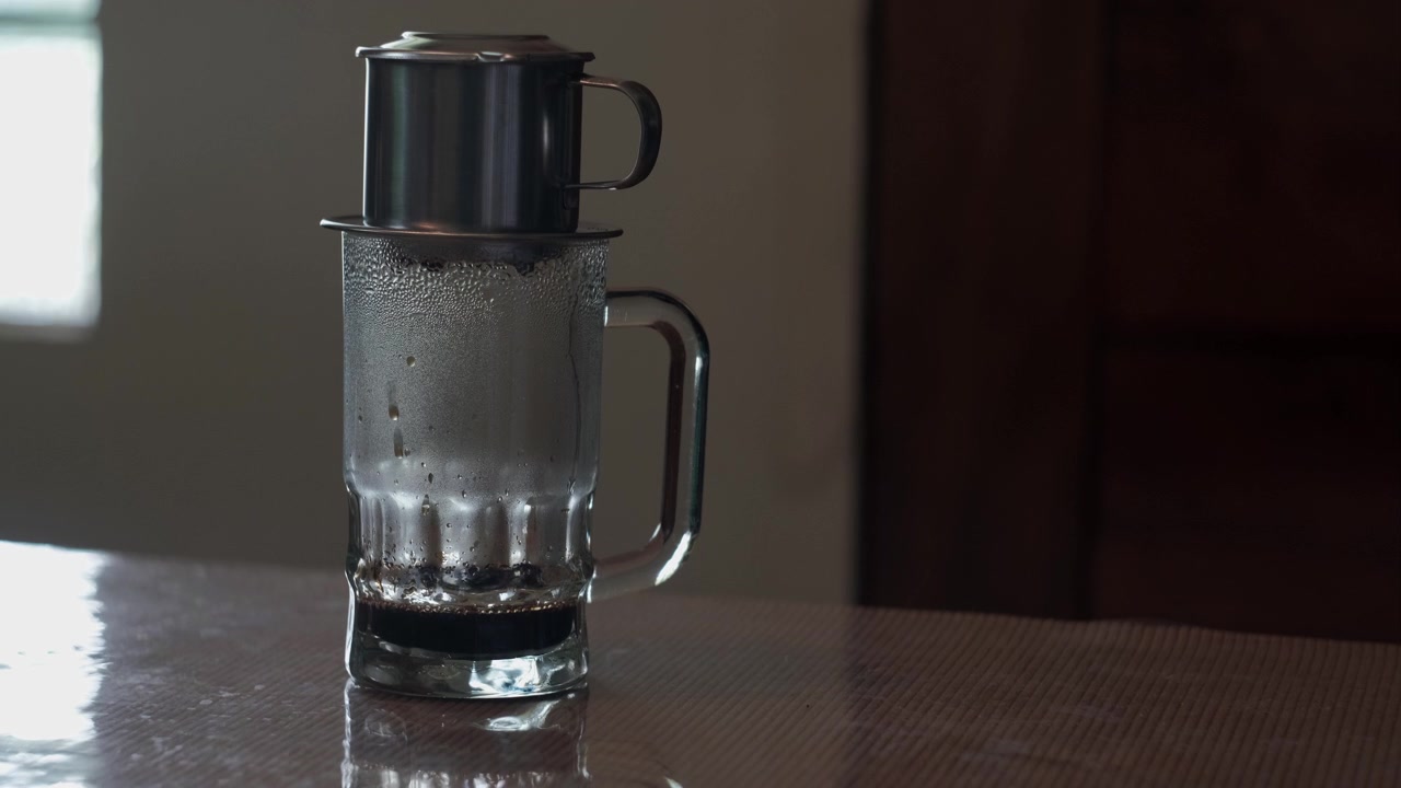 Statische Mittelaufnahme eines vietnamesischen Phin-Filters, der langsam gebrühten Kaffee in eine Glastasse auf einem Tisch in Innenräumen tropft.
