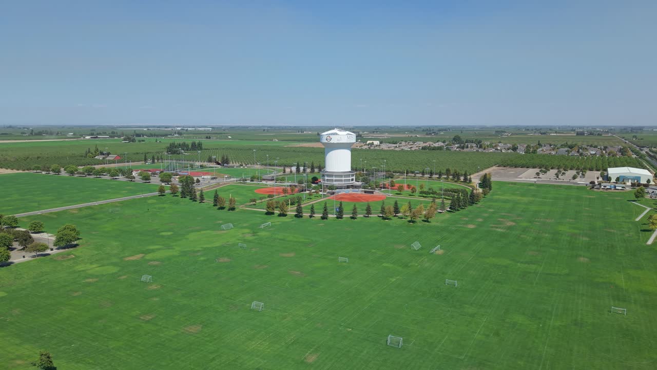 Drone lateral aerial shot showing large green sports fields with soccer goals, baseball diamonds, white water tower, parking lot and surrounding residential area in Ripon, California