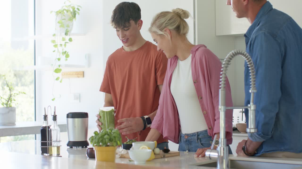 Rodina sleduje mladého člověka, jak sundává víko mixéru a podává zelené smoothie rodičům v kuchyni.