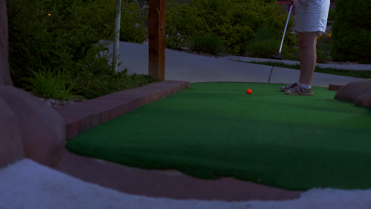 A caucasian girl mini golfing an orange golf ball hitting it towards the camera