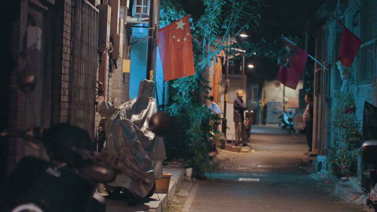 Asılı bayrak ve loş lambalarla gece sokağı, scooterlar, saksı bitkileri, çamaşırlar ve kasalarla dolu dar kentsel yol, tek başına bir yaya turkuaz-turuncu sokak lambası altında kameraya doğru yürüyor, ıslak kaldırım